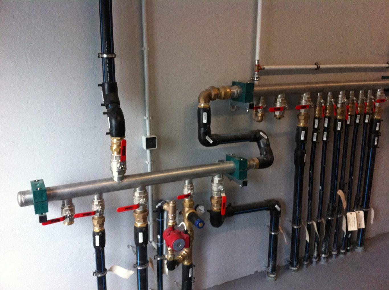 Wasserrohrinstallation mit roten Hähnen und schwarzen und metallenen Rohren an einer weißen Wand