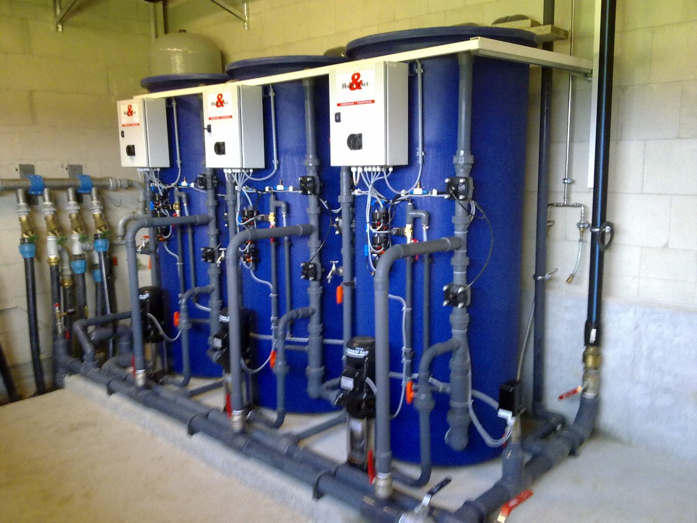 Drei blaue Wasserenthärtungen stehen nebeneinander in einem Technikraum vor einer weißen Wand.